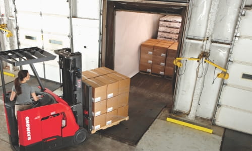 Forklift loading a pallet of cartons into a trailer at an indoor warehouse dock.