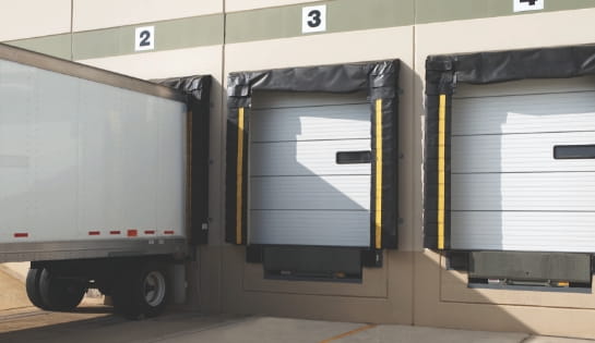 Semi-truck trailer parked at a warehouse loading dock next to closed industrial dock doors.