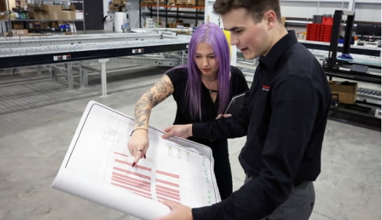 Raymond Storage Concepts project manager and engineer reviewing a racking blueprint in a warehouse with conveyors in the background.