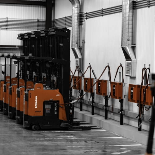 Line of Raymond reach forklifts plugged into wall-mounted battery chargers in a warehouse