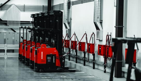 Line of Raymond reach forklifts plugged into wall-mounted battery chargers in a warehouse