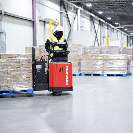 Warehouse worker driving a Raymond electric pallet jack with a loaded pallet to a staging area.