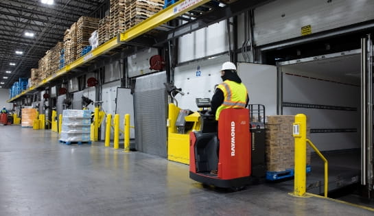 Forklift operator using a Raymond electric pallet jack to unload pallets from a trailer at a warehouse loading dock.