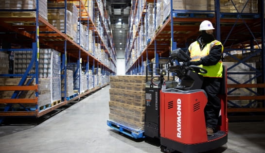 Warehouse worker wearing safety gear driving a Raymond forklift towing a pallet through a racking aisle.