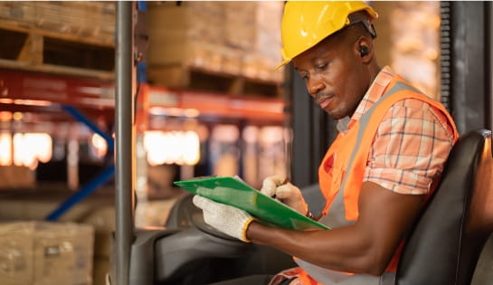 Forklift operator wearing safety gear and checking items off on a clipboard while sitting in the lift truck seat