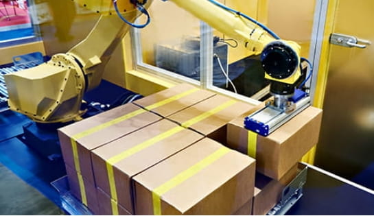 Yellow robotic arm palletizing boxes onto a wooden pallet in an automated fulfillment facility