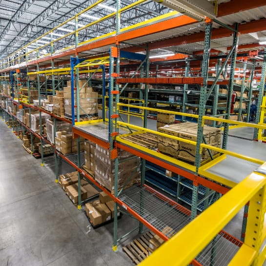 Multi-level pick module featuring cartons stored across different tiers with worker platforms and access stairs in a warehouse