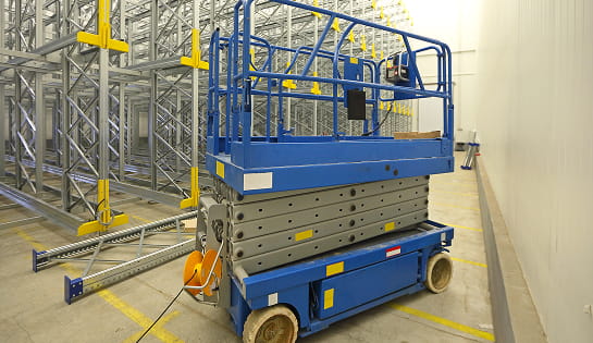 Blue scissor lift parked in a warehouse aisle with industrial metal racking in the background