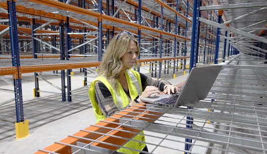 Raymond Storage Concepts account manager reviewing data on a laptop while seated on a wire deck in a warehouse racking aisle