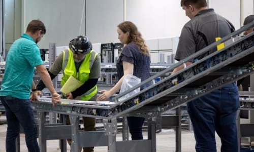 Installation lead in a hard hat and safety vest demonstrating conveyor installation progress to three Raymond Storage Concepts project managers and design engineers