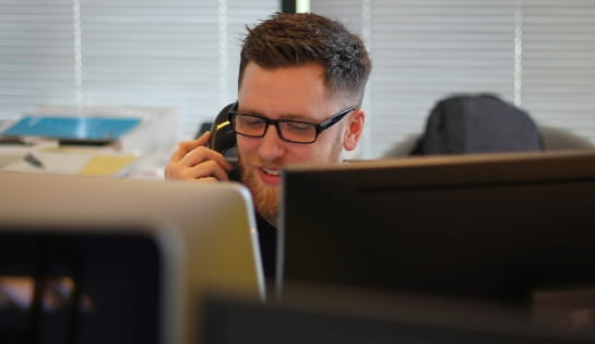 Professional man with glasses talking on the phone in an office, viewed over the top of computer monitors