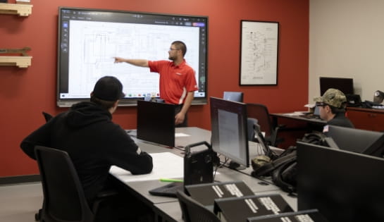 Trainer pointing at a forklift schematic on a classroom screen for technicians seated at desks with computers