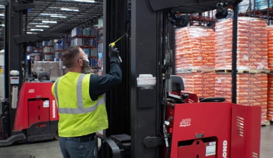 RSC technician wearing a safety vest is spraying lubricant of forklift's lift during a scheduled maintenance visit. He's in the side bay in a warehouse surrounded by other Raymond forklifts and pallets.