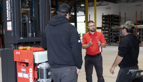 Raymond Storage Concepts trainer in a red polo talking to two technicians next to an opened forklift engine during a warehouse training session