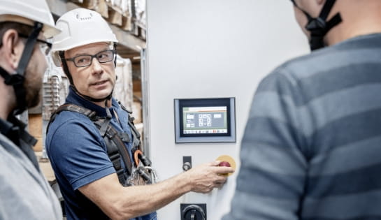 Man in a hard hat explaining equipment controllers and a monitor to two people during a demonstration