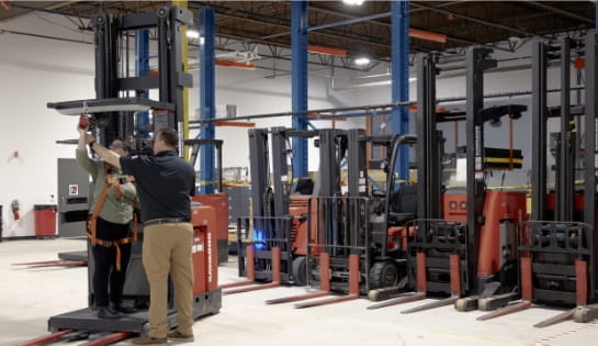 RSC trainer demonstrating a fall harness pulley to a forklift operator trainee standing on an orderpicker in a warehouse with a line of lift trucks