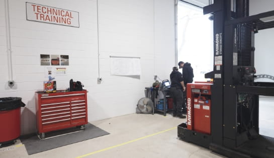 Raymond Storage Concepts technicians reviewing laptops next to an exposed forklift engine and tool drawer in the Cincinnati training facility