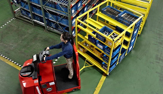 Warehouse worker driving a Raymond tugger with attached carts in a facility