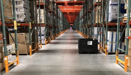 View through warehouse rack aisle pass-throughs showing fully loaded pallet racks equipped with upright guards and clear labels