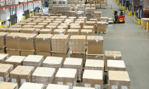 Forklift moving pallets in a busy warehouse staging area with multiple rows of storage racks